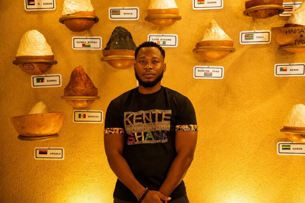 Gilbert Sepenu, founder of Vialis Travel, in a black Kente Ghana shirt in front of a wall of calabash lamps labelled with African countries